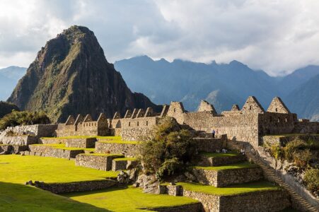 Stone ruins of Machu Picchu with terraced slopes, set against green mountains and a partly cloudy sky in the background.