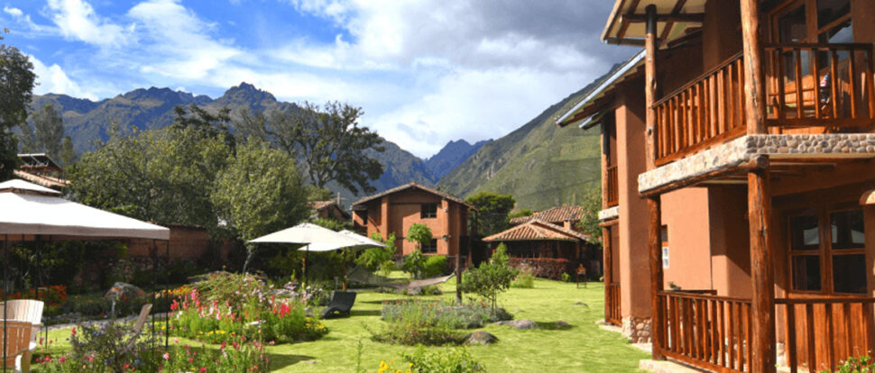 A garden with colorful flowers and umbrellas surrounded by rustic buildings, set against a backdrop of green mountains and partly cloudy skies.