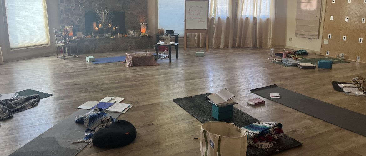 A spacious room with yoga mats, cushions, books, and papers scattered on the floor; a stone fireplace and a whiteboard are visible in the background.